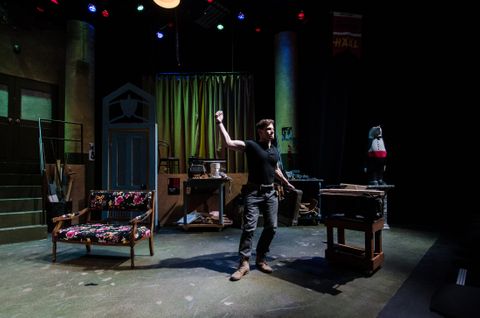 The actor playing the stage manager holds a lighter in the air as though threatening to burn the theatre down. The space is dimly lit and the actor is lit by cool, high front light.