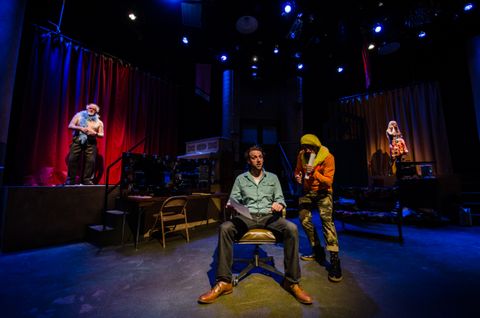 Actors are in their own specials in various parts of the space. The rest of the space is dimly lit with blue back light.